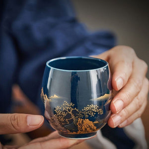 Hands holding a Kumo deep blue ceramic tea cup with gold landscape design